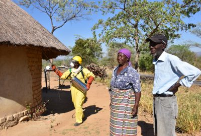 Spray campaign in Zimbabwe showing a rural home with a thatched roof on the left and a spray operator in yellow PPE and a woman with a purple head scarf and a man with a baseball cap on the right side and trees in the background and blue sky behind.