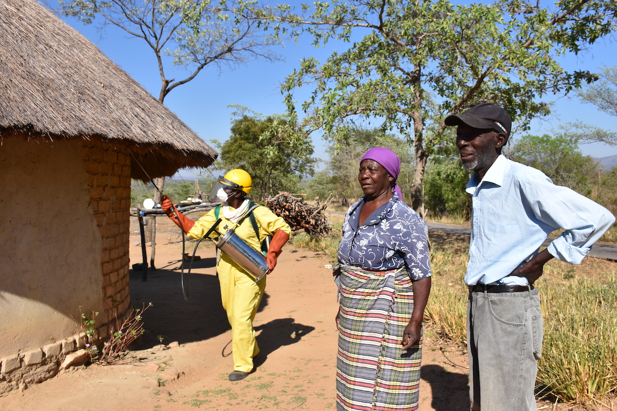 November 2018 spray campaign in Zimbabwe, PMI, 2018 Spray campaign in Zimbabwe showing a rural home with a thatched roof on the left and a spray operator in yellow PPE and a woman with a purple head scarf and a man with a baseball cap on the right side and trees in the background and blue sky behind.