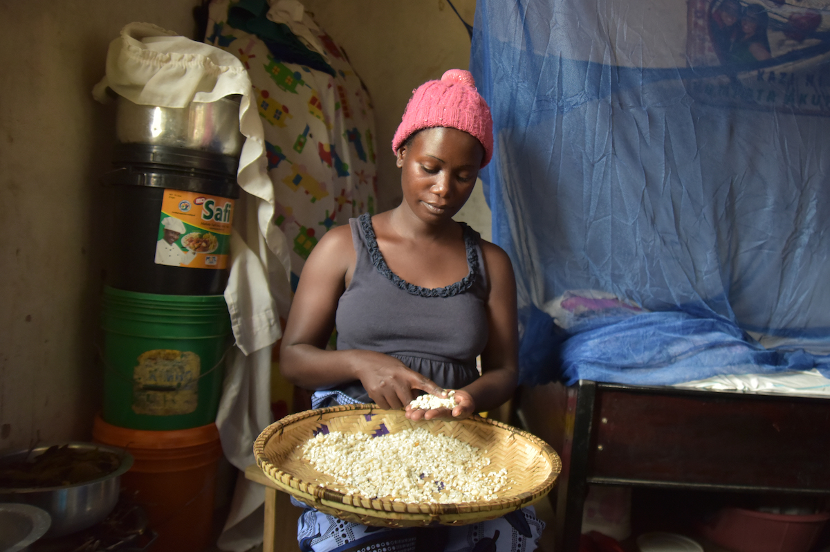 Woman holding a rice bowl and wearing a pink hat and grey shirt sitting in house with blue bednet behind her.