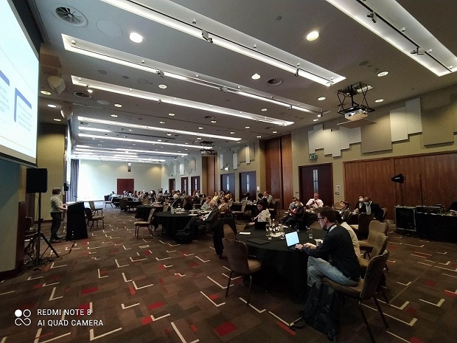 Photograph from the I2I May 2022 Convening showing a large conference room with people sitting at round tables with a speaker presenting at a podium and a presentation on a screen behind the podium.