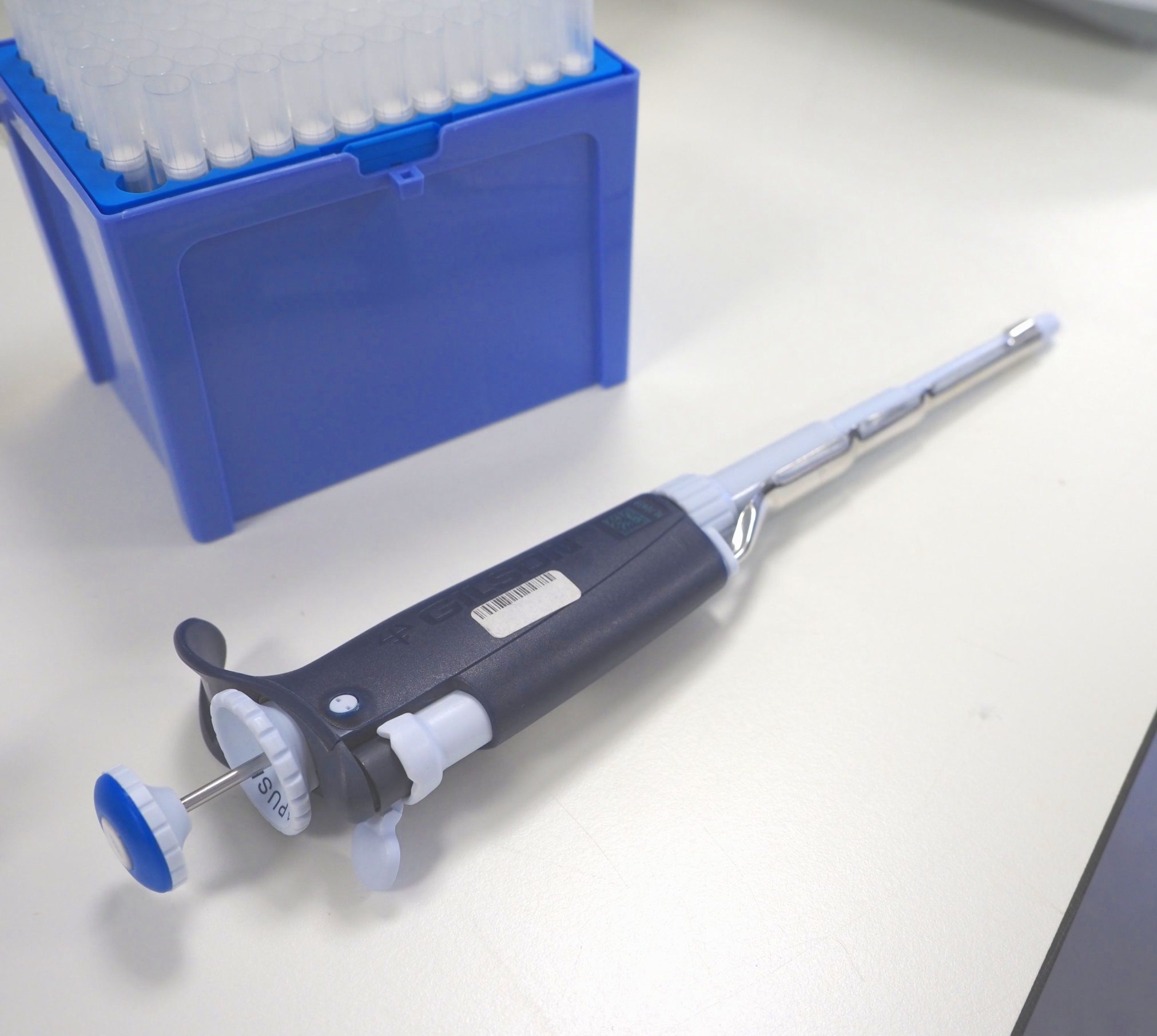 Open blue pipette tip box in upper left corner of image with dark blue and white pipette without tip lying on a white lab table