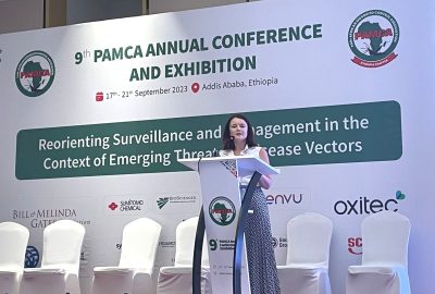 A woman with brown short hair, wearing a white short sleeve shirt and blue trousers standing at a white podium with the PAMCA logo on it on a stage in front of a PAMCA banner and a row of white chairs.