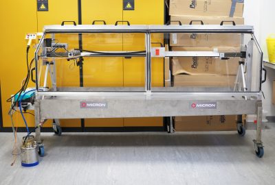 Image of the Track Sprayer. a horizontal, rectangular metal structure with glass doors on all sides with a canister at the base on the left side. Bright yellow cabinets are behind the Track Sprayer.