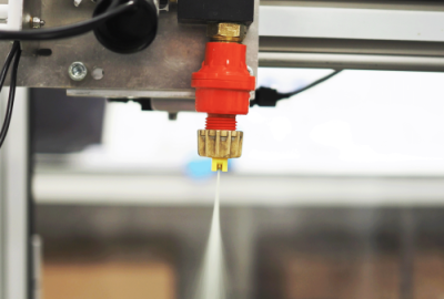 Image of the Micron Horizontal Track Sprayer spray nozzle in action. In the centre of the image is a red and gold nozzle on a silver and black metal track that is horizontal across the image. A fine stream of liquid is spraying downward from the nozzle. Behind the nozzle is a glass window with a silver metal frame.