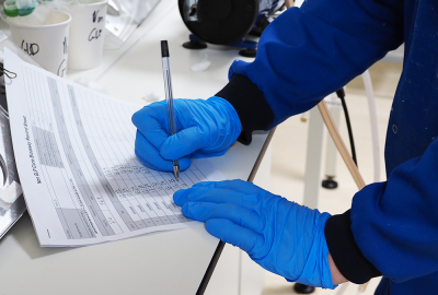 Picture of two hands in latex blue gloves of person wearing a royal blue lab coat. The left hand is holding a data collection sheet in place on a lab counter on the left of the image. The right hand is using a black pen to write data entries. Behind the data collection sheet is a white cup holding tubes with green rubber bands around the top of the tubes.